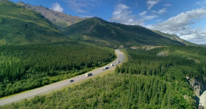 Alaska. Aerial 4K. Drone flight over the highway and a forest in a mountains. Camera moves above the tops of the trees. On the road there are white RVs and a passenger car driving in same direction.