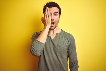 Young handsome man wearing casual t-shirt standing over isolated yellow background covering one eye with hand, confident smile on face and surprise emotion.