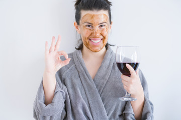 Beautiful woman wearing cosmetic facial mask as skincare treatment drinking glass of wine doing ok sign with fingers, excellent symbol