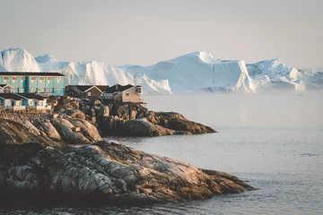 Luftaufnahme der arktischen Stadt Ilulissat, Grönland bei Sonnenaufgang Sonnenuntergang mit Nebel. Bunte Häuser im Zentrum der Stadt mit Eisbergen im Hintergrund im Sommer an einem sonnigen Tag mit orangefarbenem Rosa © Mathias