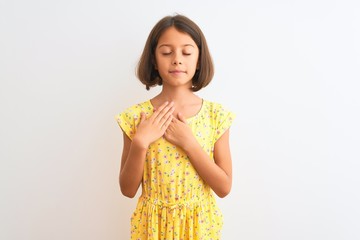Young beautiful child girl wearing yellow floral dress standing over isolated white background smiling with hands on chest with closed eyes and grateful gesture on face. Health concept.