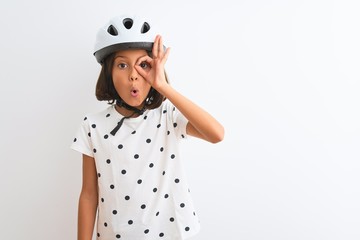 Beautiful child girl wearing security bike helmet standing over isolated white background doing ok gesture shocked with surprised face, eye looking through fingers. Unbelieving expression.