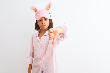 Beautiful child girl wearing sleep mask and pajama standing over isolated white background looking unhappy and angry showing rejection and negative with thumbs down gesture. Bad expression.
