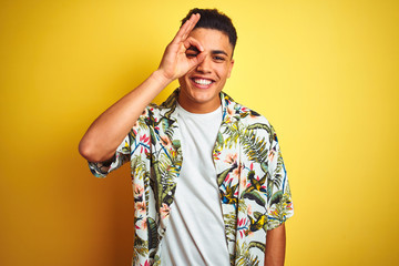 Young brazilian man on vacation wearing summer floral shirt over isolated yellow background doing ok gesture with hand smiling, eye looking through fingers with happy face.