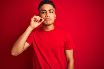 Young brazilian man wearing t-shirt standing over isolated red background mouth and lips shut as...