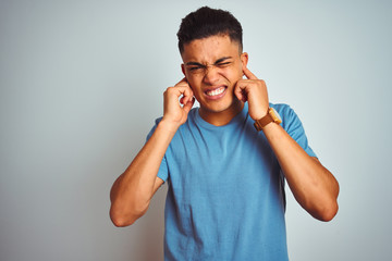 Young brazilian man wearing blue t-shirt standing over isolated white background covering ears with fingers with annoyed expression for the noise of loud music. Deaf concept.