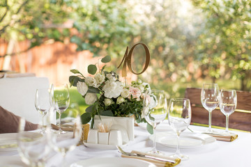 Wedding table setting decorated with fresh flowers in a brass vase. Wedding floristry. Banquet table for guests outdoors with a view of green nature. Bouquet with roses, eustoma and eucalyptus leaves