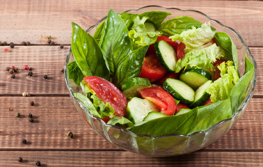 Salad of greens, tomato and cucumbers in a glass plate on an old wooden background.