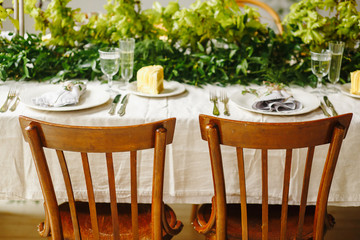 Wedding table with chairs and plates, forks, spoons, knifes on cloth decorated with greenery bouqet