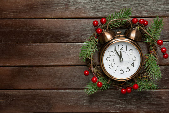 Traditional Handmade Christmas Wreath With Green Fir Tree Branches Holly Berries Clock Showing Five Minutes To Midnight On Dark Wood Background. New Years Countdown Concept