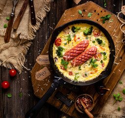 overhead shot of homemade tasty baked omelette or frittata with two sausages, broccoli, cherry tomatoes, basil, melted cheese in black skillet on wooden board on rustic table with sackcloth