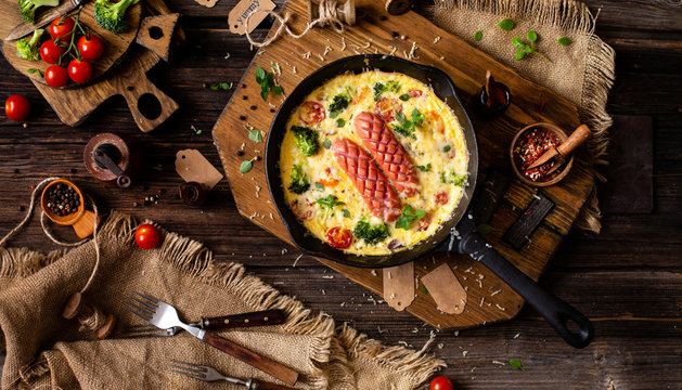 overhead shot of homemade tasty baked omelette or frittata with two sausages, broccoli, cherry tomatoes, basil, melted cheese in black skillet on wooden board on rustic table with sackcloth