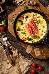 overhead shot of homemade tasty baked omelette or frittata with two sausages, broccoli, cherry tomatoes, basil, melted cheese in black skillet on wooden board on rustic table with sackcloth