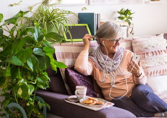 Fototapeta na wymiar One senior people, pretty grandmother with gray hair and eyeglasses rests on the sofa with romantic pillows and talks with her cellphone. Retirement in relaxation. Books and tablet on background