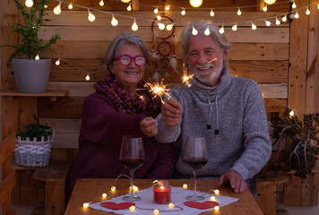 A smiling senior couple with sparkles sitting outdoor in a romantic corner made by wood. Yellow lights. Two red wineglasses to celebrate a positive moment