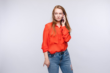 Front view of pretty girl wearing red blouse and jeans talking by phone on isolated background. Female looking aside and thinking. Concept of communication and conversation.