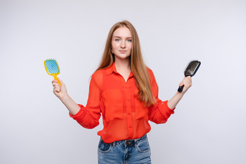 Front view of young woman keeping in hands two brushes and choosing hair care. Longhaired girl...
