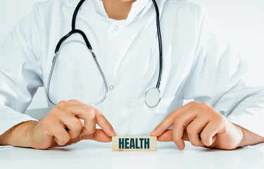 The doctor holds a block with the word HEALTH in his hand. Medical and pharmaceutical concept. A man wearing a white coat and stethoscope heals.