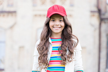 This girl is hipster. Happy little hipster with long curly hair wearing baseball cap outdoor. Cute small hipster smiling with fashionable look. Adorable child in hipster style