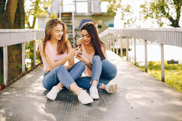 two beautiful and bright friends in pink t-shirts and blue jeans sitting in the sunny summer park and use the phone
