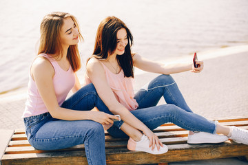 two beautiful and bright friends in pink t-shirts and blue jeans sitting in the sunny summer city and use the phone with headphones