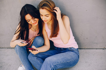 two beautiful and bright friends in pink t-shirts and blue jeans sitting in the sunny summer city near gray wall