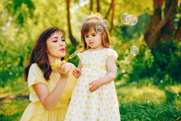 in a summer solar park near green trees, mom walks in a yellow dress and her little pretty girl and...
