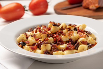 Homemade italian gnocchi with dry tomato, bacon and parmesan cheese in a white plate on white wooden table.  Soft light