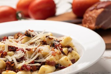 Homemade italian gnocchi with dry tomato, bacon and parmesan cheese in a white plate on white wooden table.  Soft light