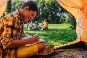 selective focus of happy boy writing in notebook in camp