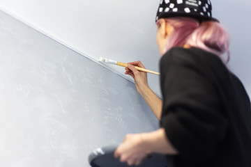 Back view of young girl with pink hair, painting wall with brush and white color, in apartment.