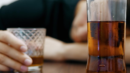 In the background a blurred young drunk man with a glass of whiskey in his hand is sleeping on the table, a half empty bottle of whiskey in the foreground in focus