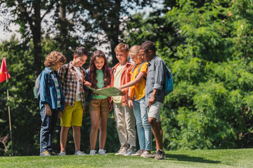 happy group of multicultural kids looking at map near trees in park