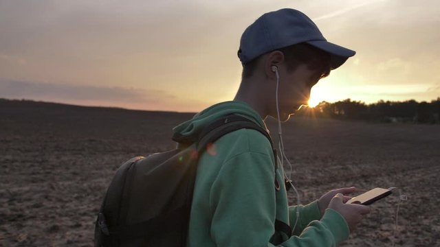 boy with backpack walks outdoors at sunset and uses the phone, listening to music