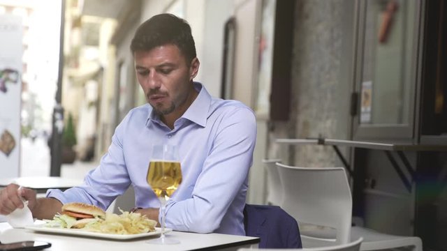 Funny close up of hungry starving handsome and young businessman eating devouring a big hamburger during lunch break. Eating chips and drinking a beer.