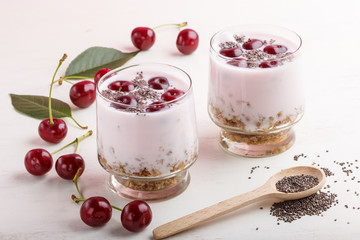 Yoghurt with cherries, chia seeds and granola in glass with wooden spoon on white wooden background. side  view.