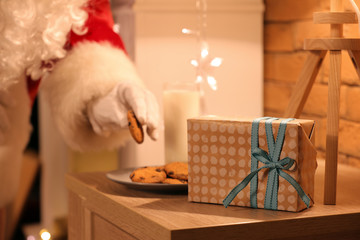 Santa Claus taking cookie from table with Christmas gift in room