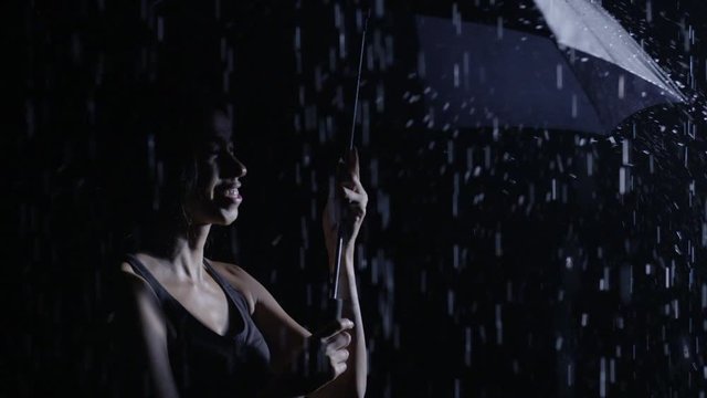 CU Young mixed-race woman twirling umbrella with rain falling at night in slow motion. Dark, high contrast footage recorded at 120fps.