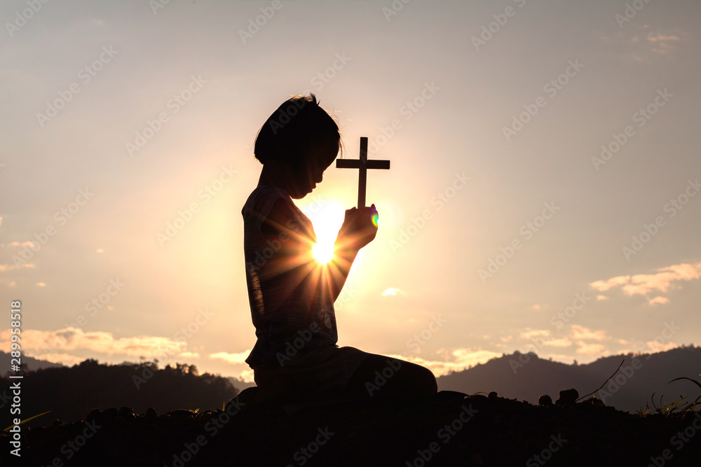 Wall mural Silhouette child praying to the GOD while holding a crucifix symbol with bright sunbeam on the sky