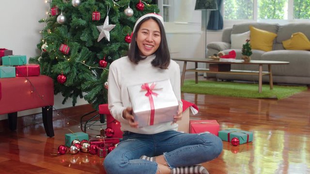 Asian women celebrate Christmas festival. Female teen wear sweater relax happy hold gift smiling near Christmas tree enjoy xmas winter holidays together in living room at home. Slow motion shot.