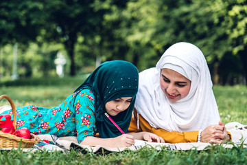 Portrait of happy muslim mother and little muslim girls child learning together with hijab dress smiling and enjoy relax reading and write a book in summer park.Education concept