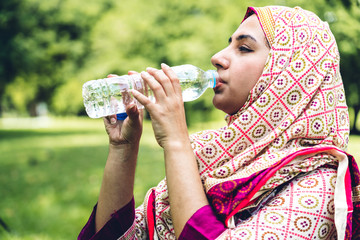 Portrait of happy arabic muslim woman with hijab dress drinking water from a bottle while relaxing and feeling fresh on green natural background at summer green park. Healthy lifestyle concept