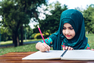 Portrait of happy little muslim girls child with hijab dress smiling and enjoy relax reading and write a book in summer park.Education concept