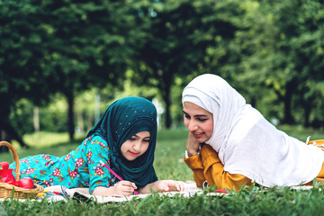 Portrait of happy muslim mother and little muslim girls child learning together with hijab dress smiling and enjoy relax reading and write a book in summer park.Education concept