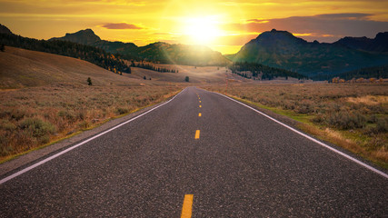 Long empty stretch of a two-lane highway heading toward a golden sunrise over a mountain pass on the horizon. Conceptual for freedom, enjoying the journey, and the promise of a new day.