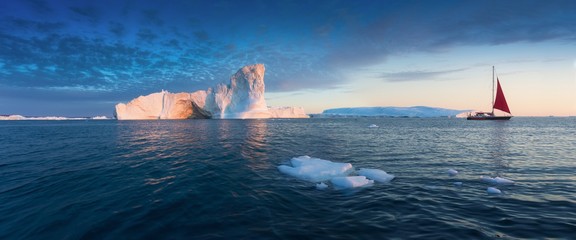 Little red sailboat cruising among floating icebergs in Disko Bay glacier during midnight sun season of polar summer. Ilulissat, Greenland. Unesco world heritage © Michal