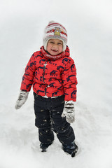 A child in a red jacket playing in the snow. The cute boy walks in the winter on the street. The kid laughs and looks at the camera. Happy childhood, fun weekend in nature. It snowed,winter is coming.