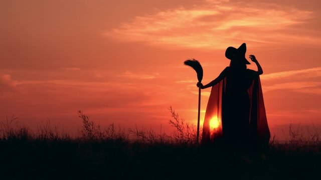 woman in halloween costume sorceress holding besom posing on the hill warm sunrise with yellow sun in the sky
