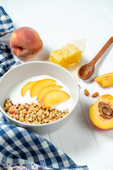 white wheat granola with low-fat yogurt in a white bowl in a composition with a spoon, honeycombs and peach on white wooden background. Healthy breakfast food. Making breakfast. Gluten free