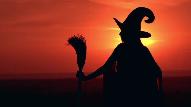 profile woman in halloween costume sorceress holding besom waiting posing on the hill sky with yellow sun
